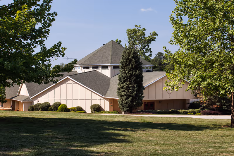 A beige senior living facility building with a multi-faceted roof on a green lawn, framed by trees.