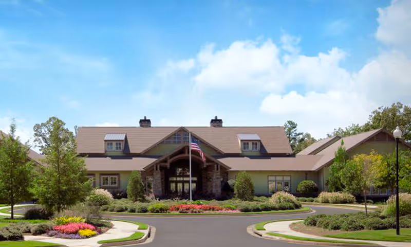 Front exterior view of a large, single-story building with a peaked roof, surrounded by landscaped gardens and trees under a blue sky with some clouds. An American flag is displayed on a flagpole in front of the entrance.