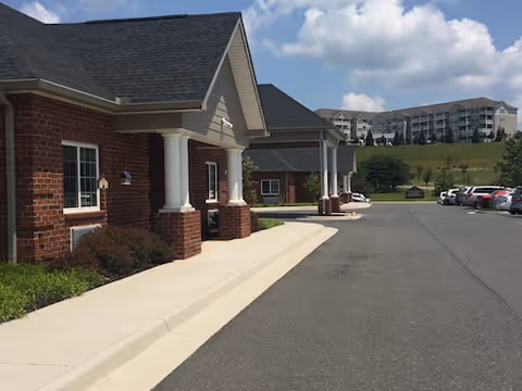 Front exterior entrance of a brick health and rehabilitation center with a covered drive, parking area, and multi-story buildings on a grassy hill under a blue sky.