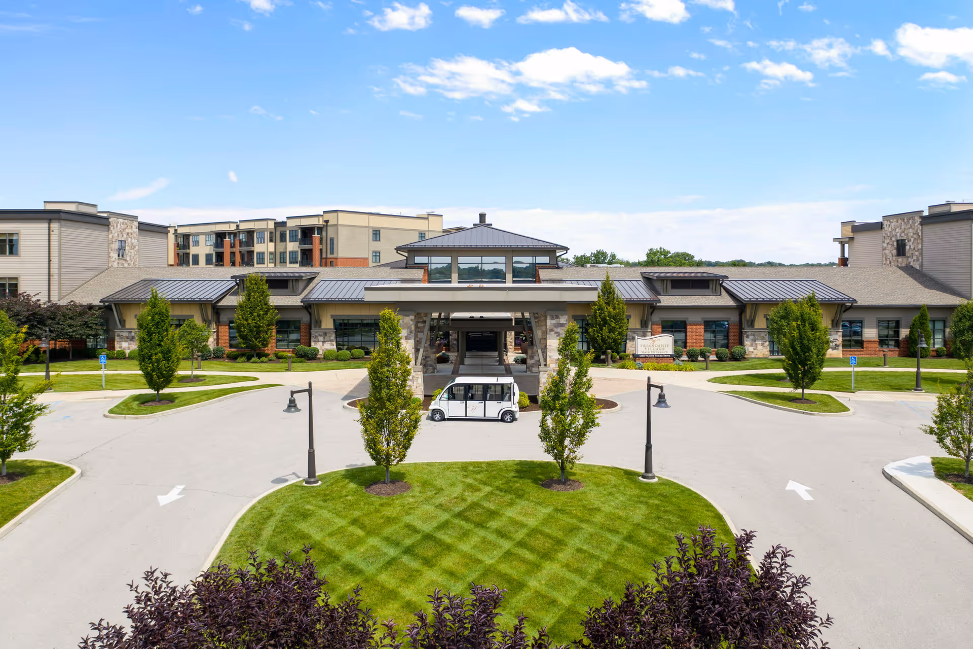 Wide view of Friendship Village Sunset Hills front entrance, curved driveway, green lawns, and trees.
