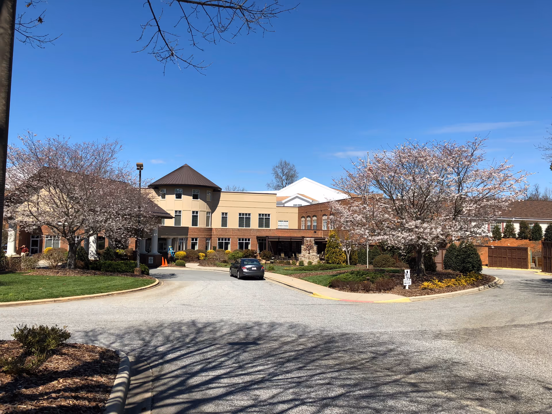 Exterior view of a senior living facility named Trinity Oaks on a clear sunny day. The building has a mix of beige and brick walls with multiple windows and a round tower-like structure. There are blooming trees with pink flowers and well-maintained landscaping around the driveway where a car is parked.