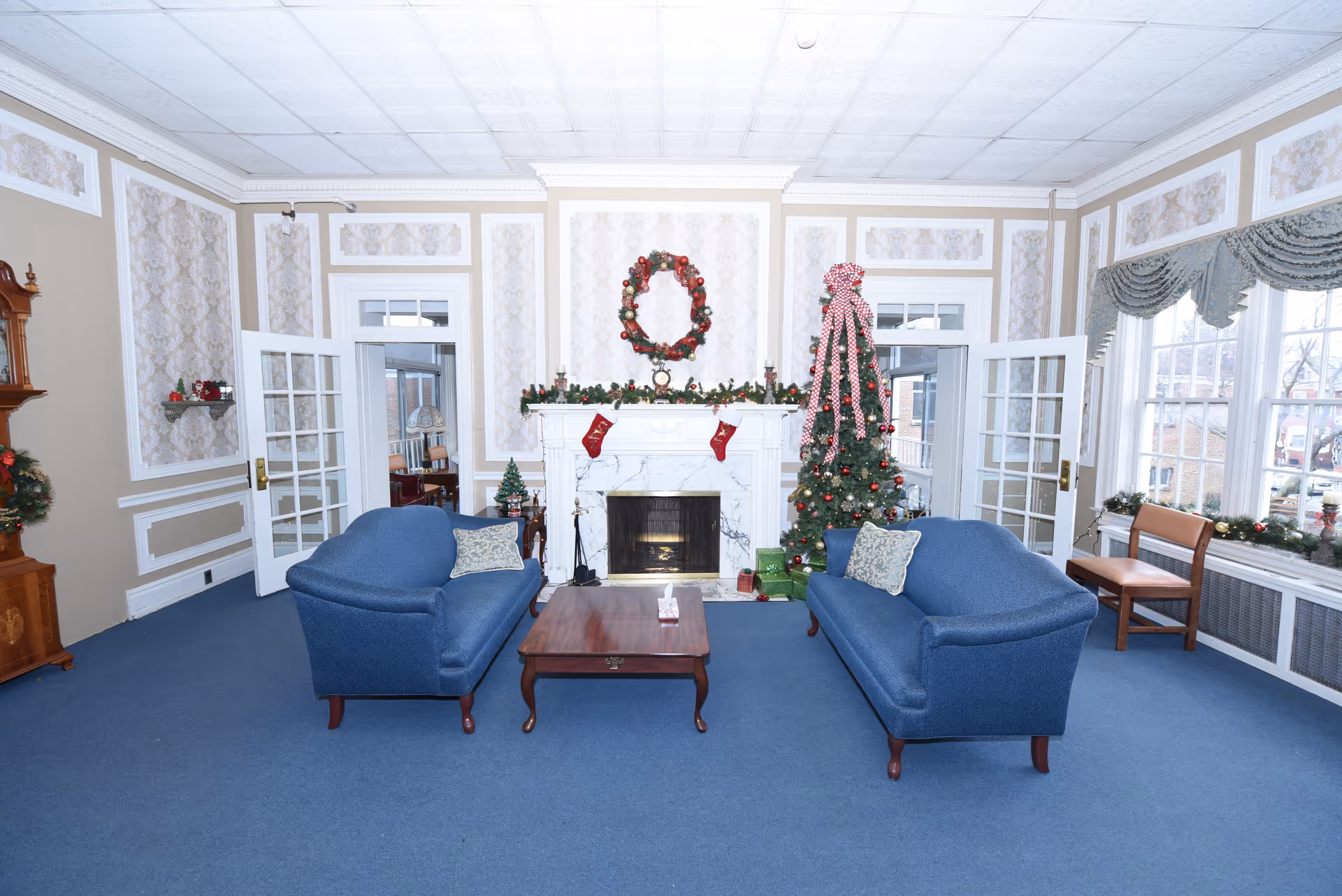 A cozy living room decorated for Christmas with two blue upholstered sofas facing each other across a wooden coffee table. A white marble fireplace is adorned with a festive wreath, garland, and red stockings. To the right of the fireplace stands a decorated Christmas tree with red ornaments and a large red and white bow on top. The room features large windows with draped curtains, patterned wallpaper, and blue carpeted flooring.
