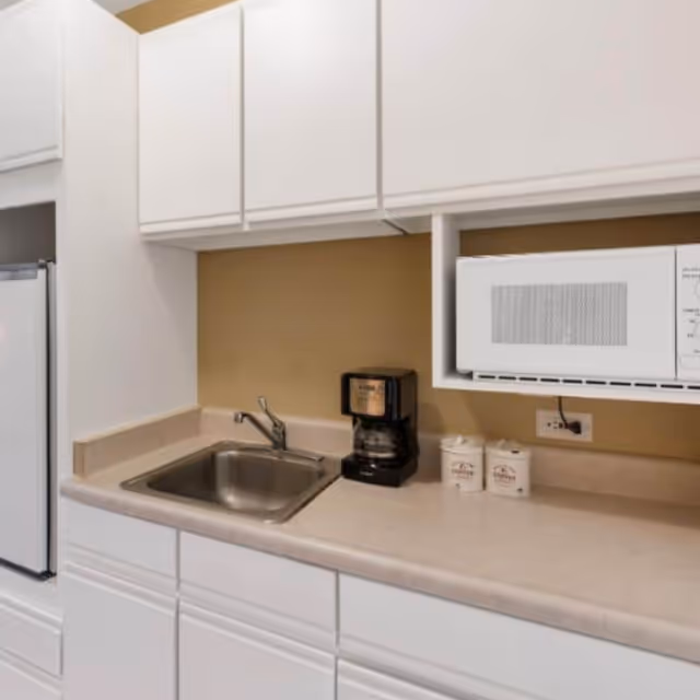 White kitchen with sink, coffee maker, microwave, and beige countertops in a senior living facility.