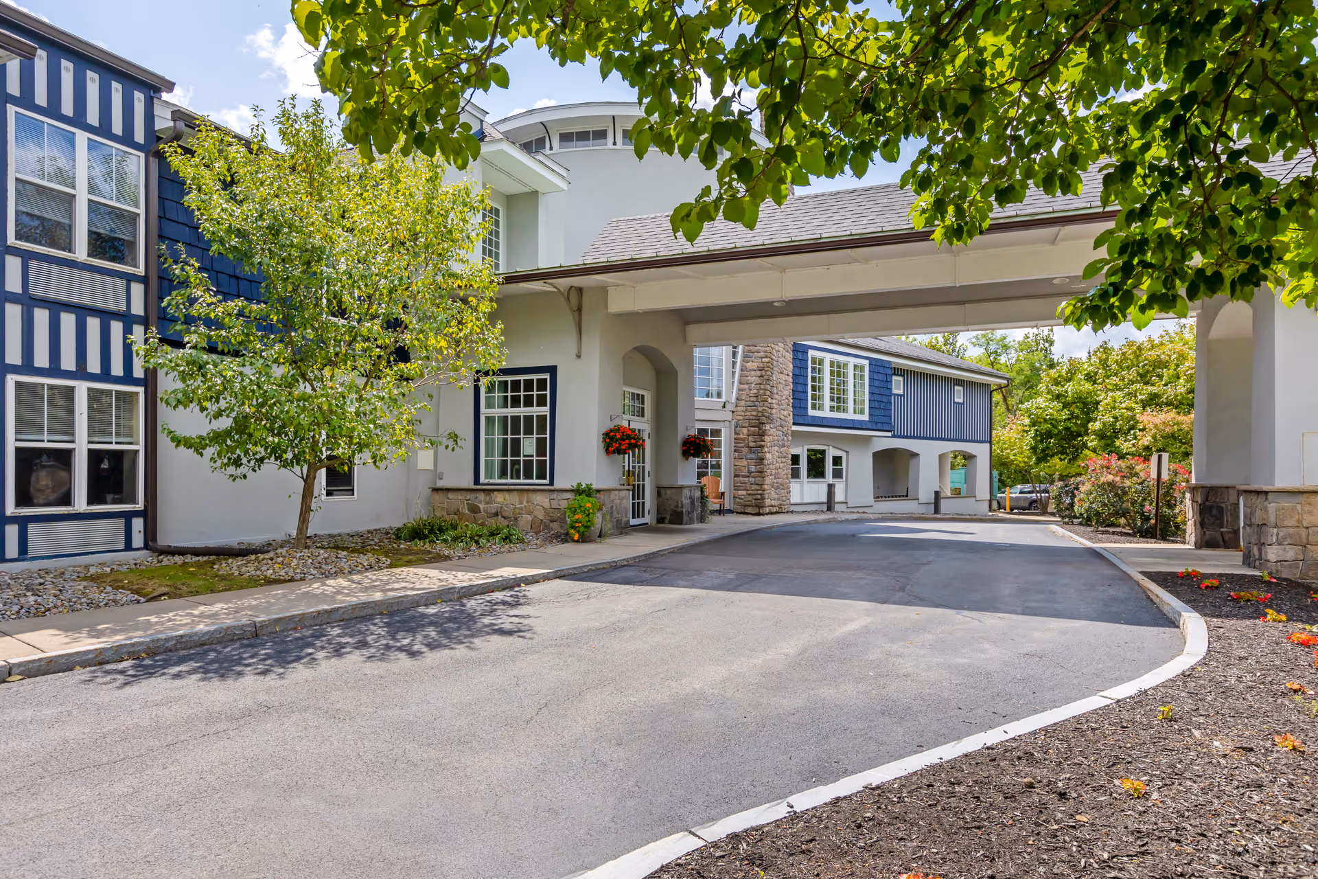 Covered driveway and front entrance of a blue-and-white senior living building with trees and landscaping.