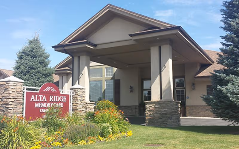 Exterior front entrance of Alta Ridge Memory Care of Sandy facility with a covered driveway, stone pillars, large windows, and landscaped garden with flowers and shrubs.
