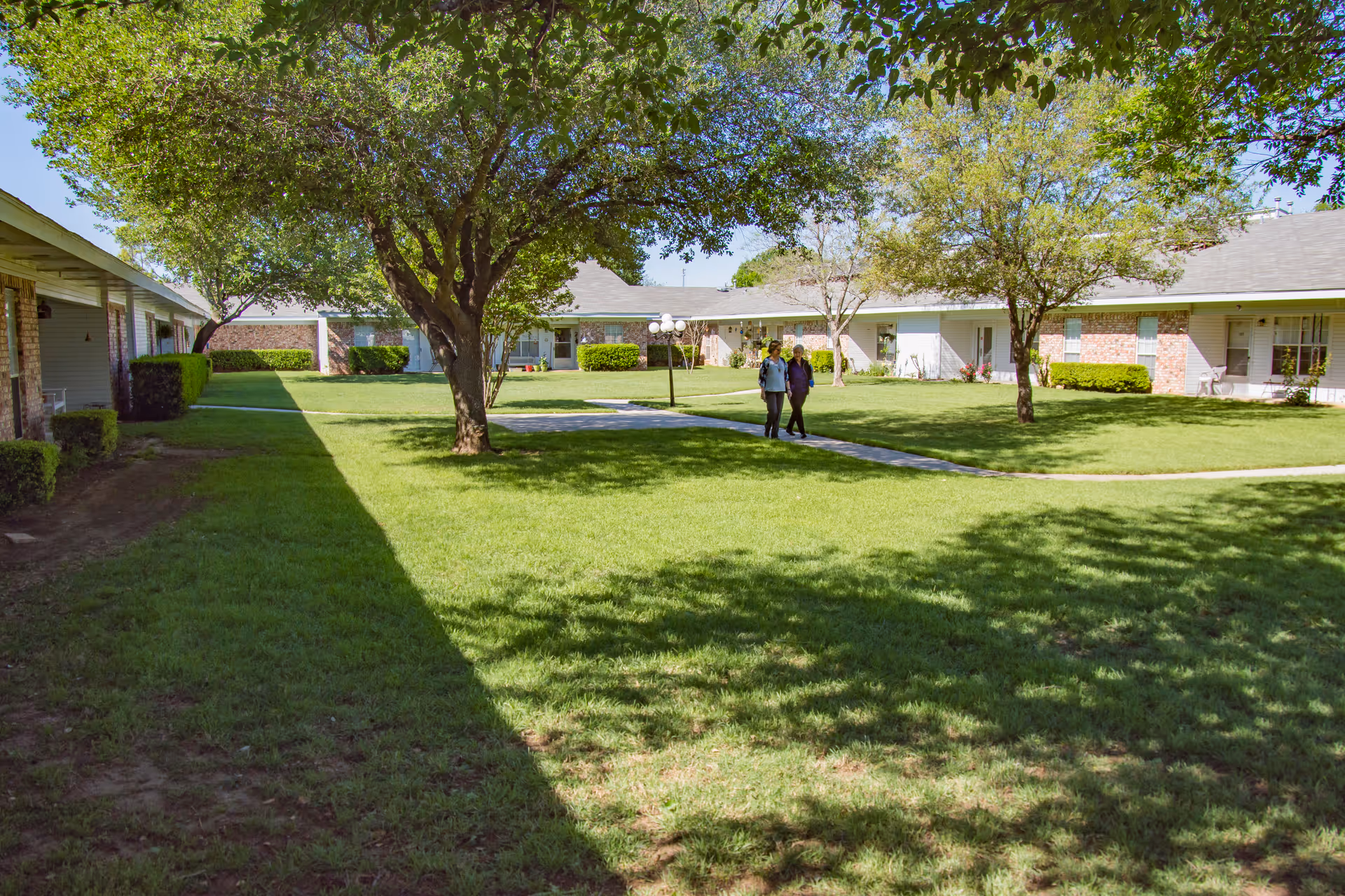 A sunny outdoor courtyard area at Vicksburg Retirement Community with green grass, several trees providing shade, and a paved walkway. Two people are walking along the path. Surrounding the courtyard are single-story buildings with brick and siding exteriors.