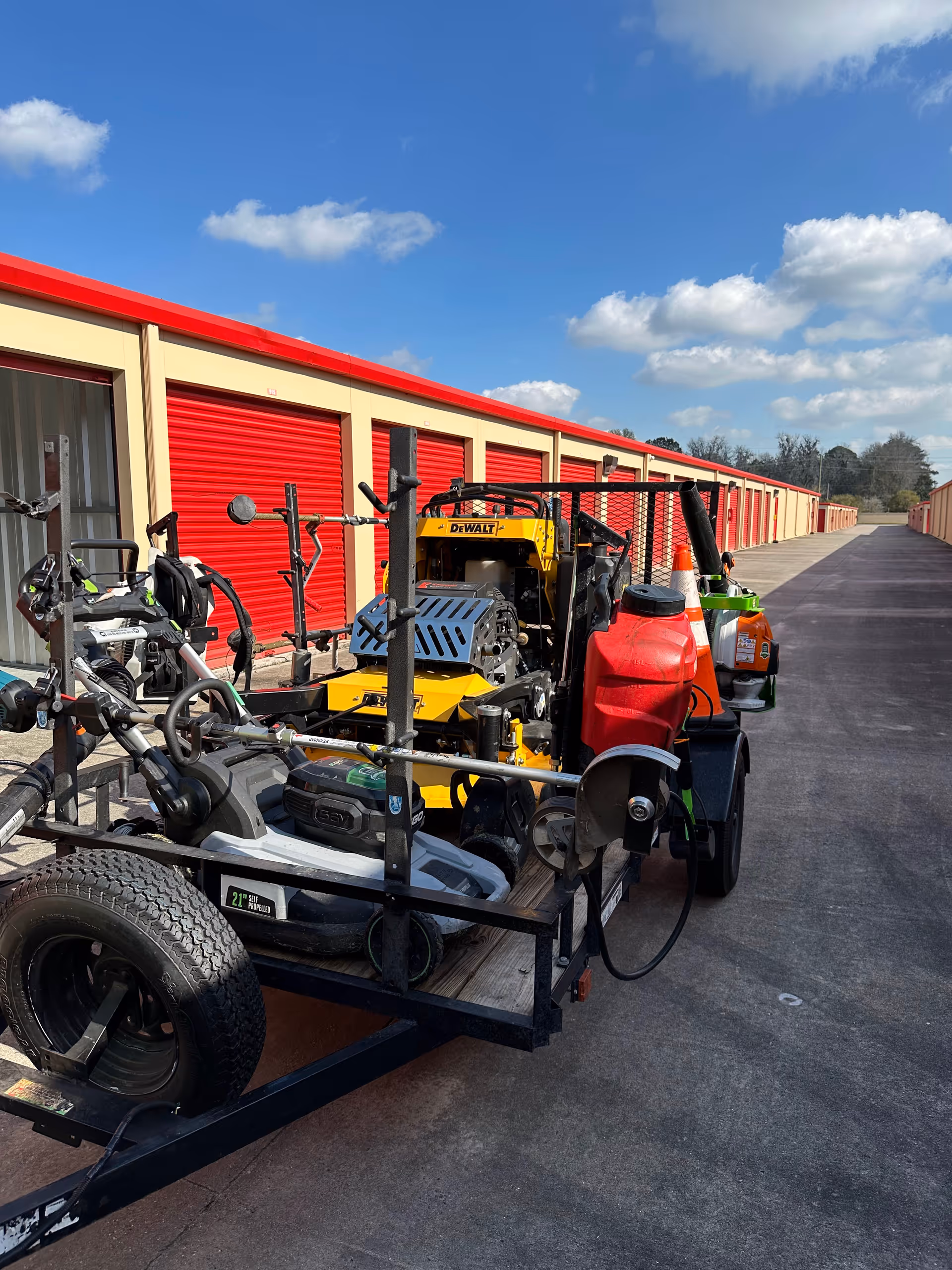 Outdoor storage lot with a trailer carrying lawn and maintenance equipment beside red storage units.