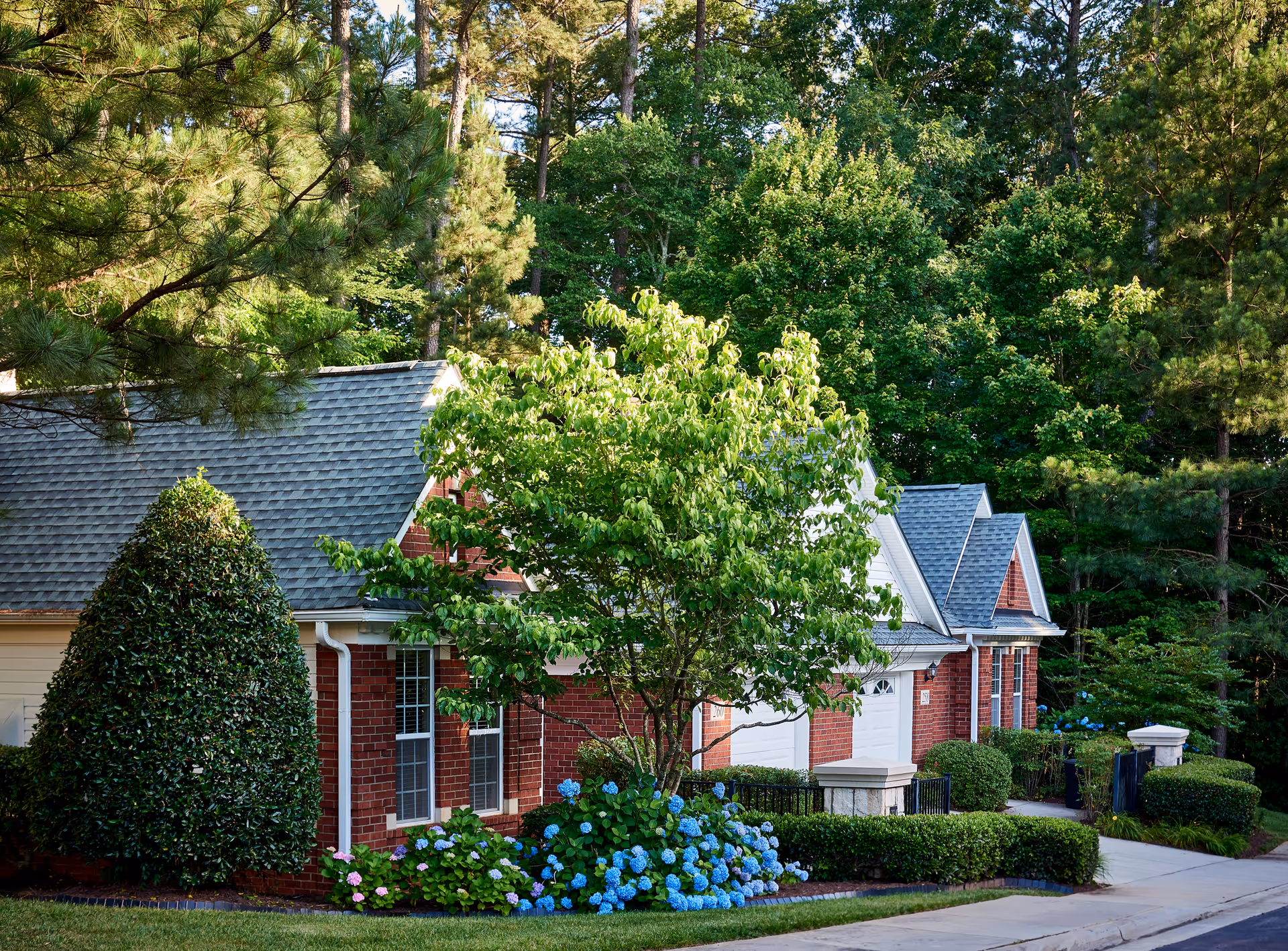 Exterior view of a residential-style building with red brick walls and gray shingled roofs, surrounded by lush green trees, bushes, and blooming blue and pink hydrangea flowers along a paved driveway.