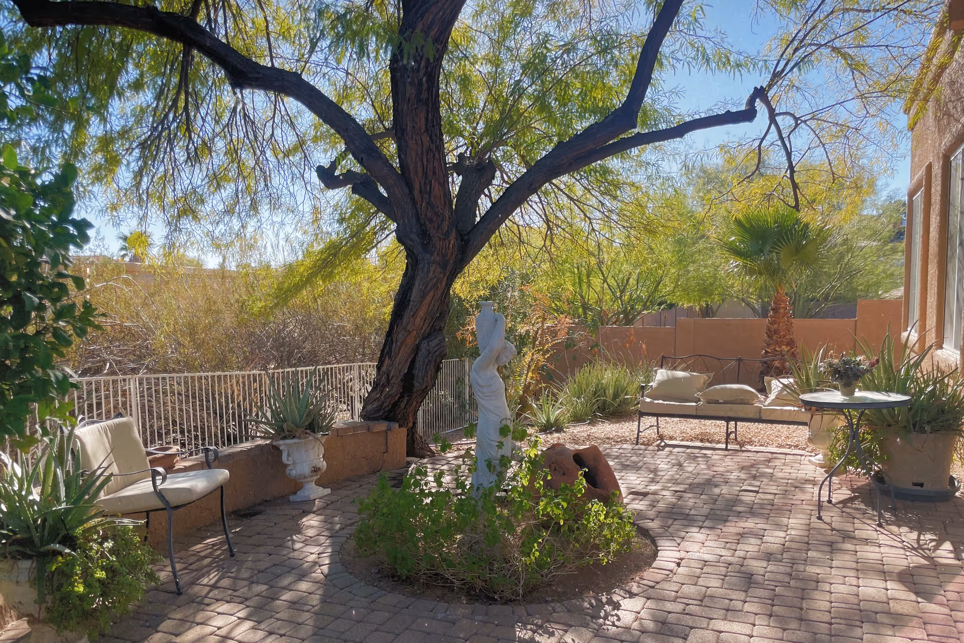 Sunny outdoor courtyard with brick patio, seating, plants, and a central statue.