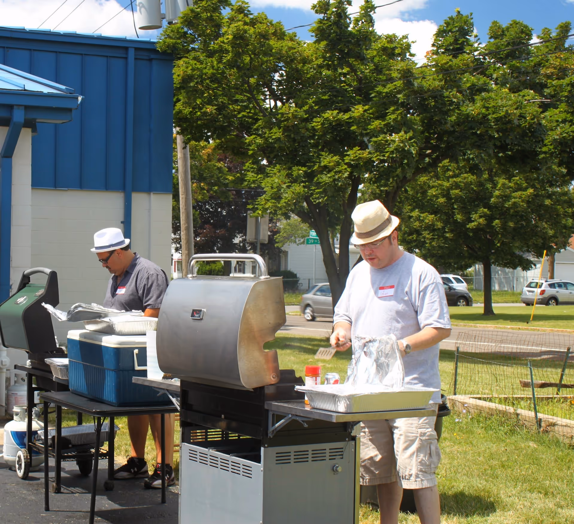 Two men grill outdoors beside a blue building with trees and lawn.
