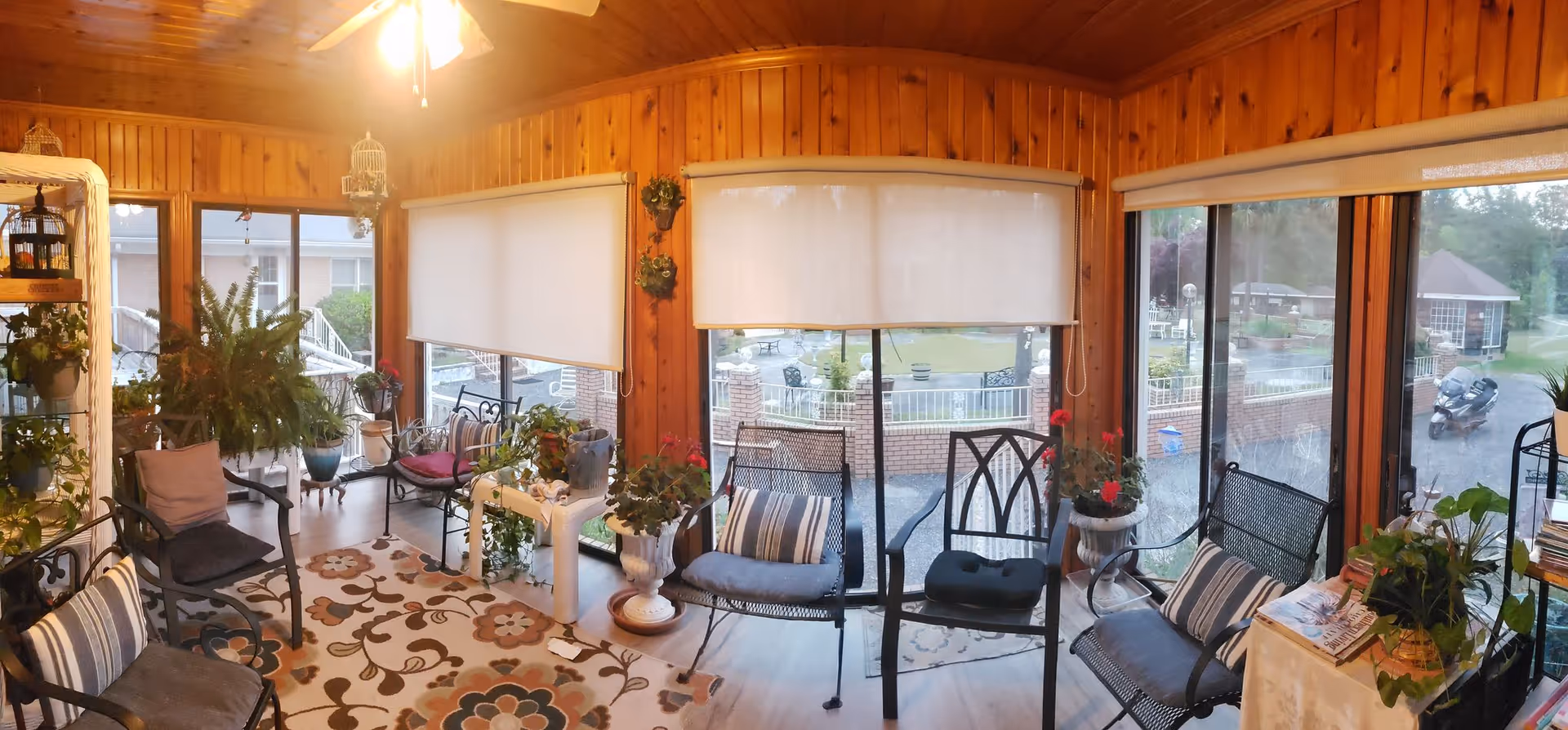 Sunlit enclosed porch with wood paneling, seating, and abundant potted plants.