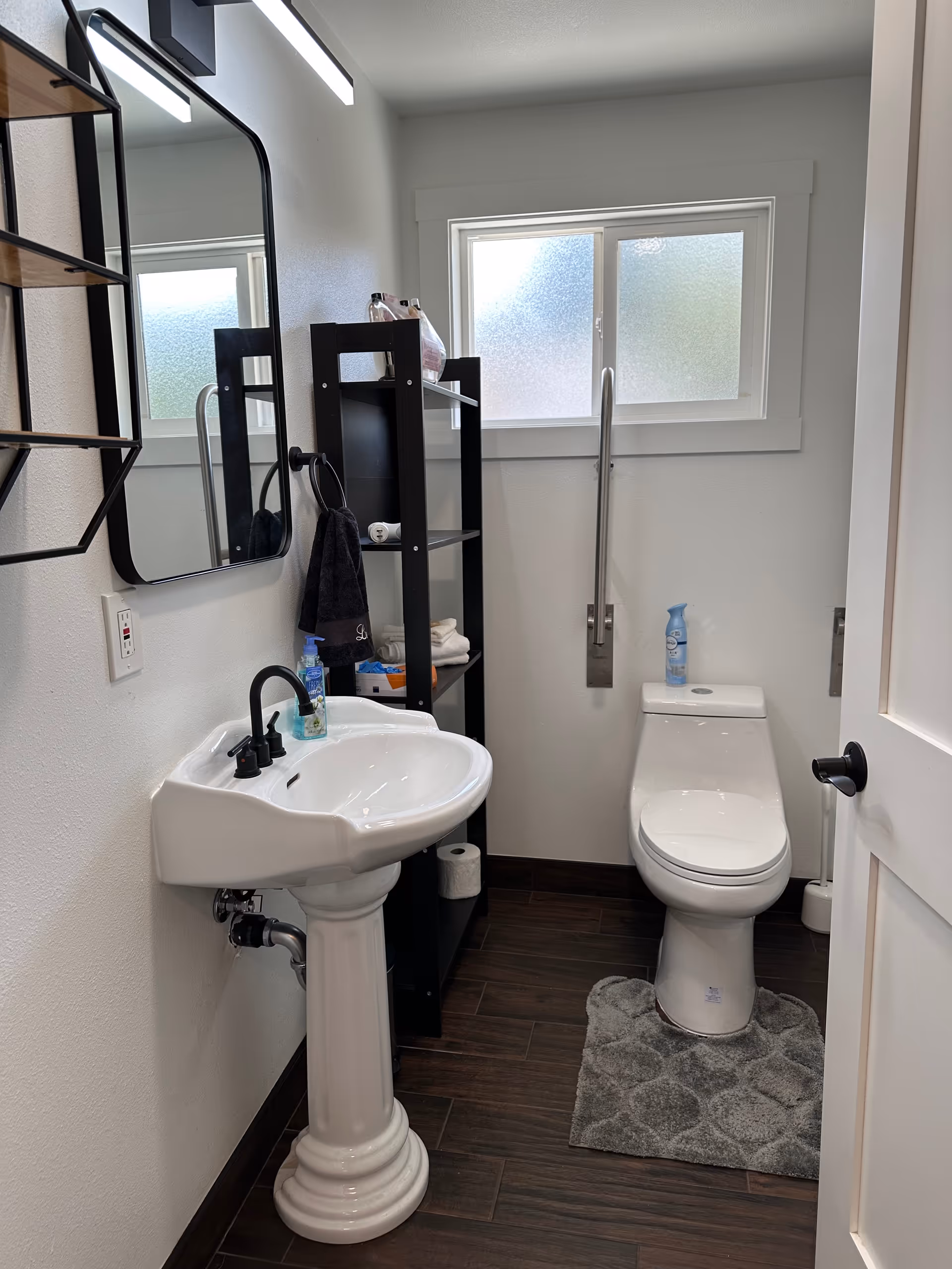 Compact bathroom featuring pedestal sink, toilet, black shelving, and frosted window.