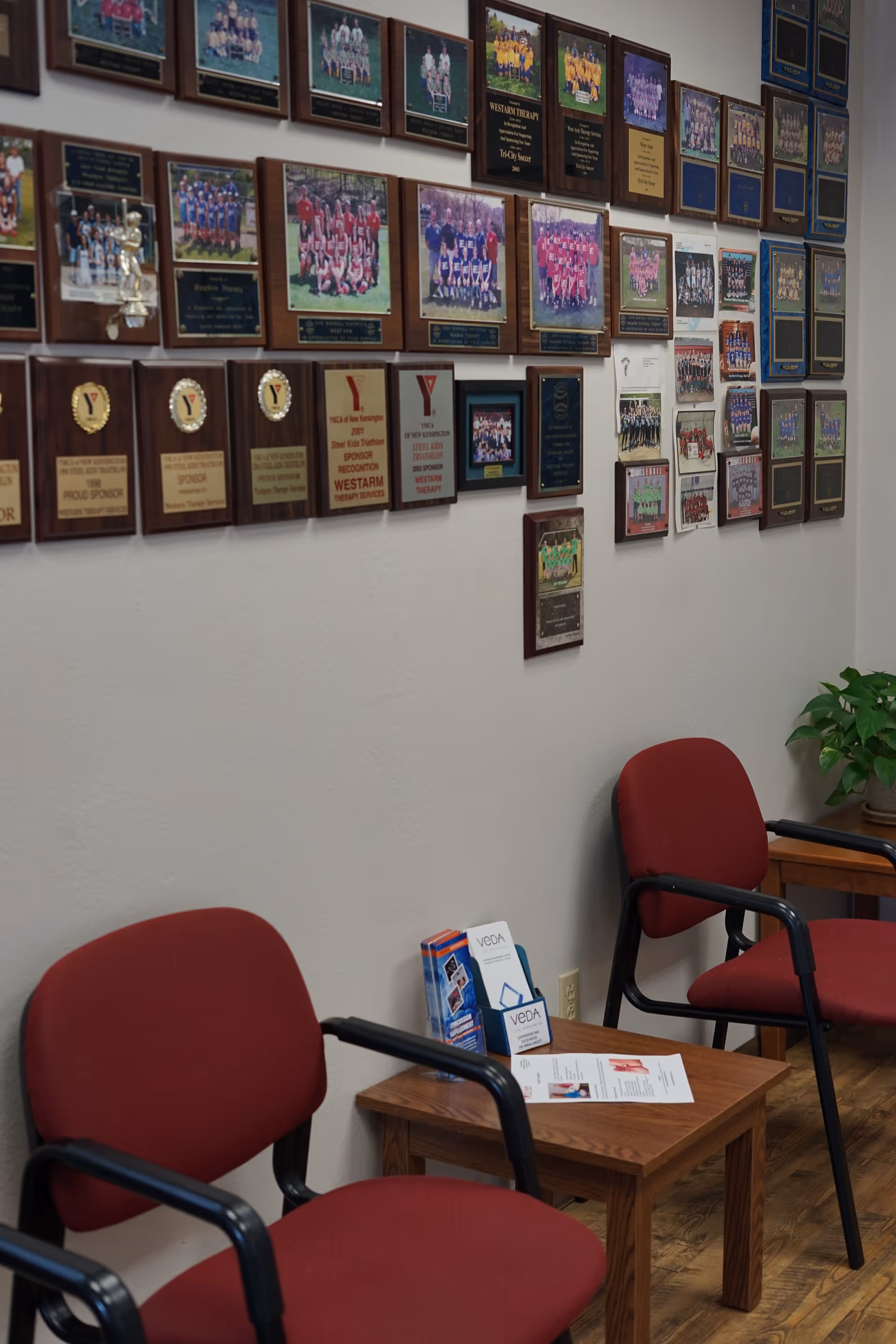 Interior lounge with red chairs and a table beside a wall covered in plaques and photographs.