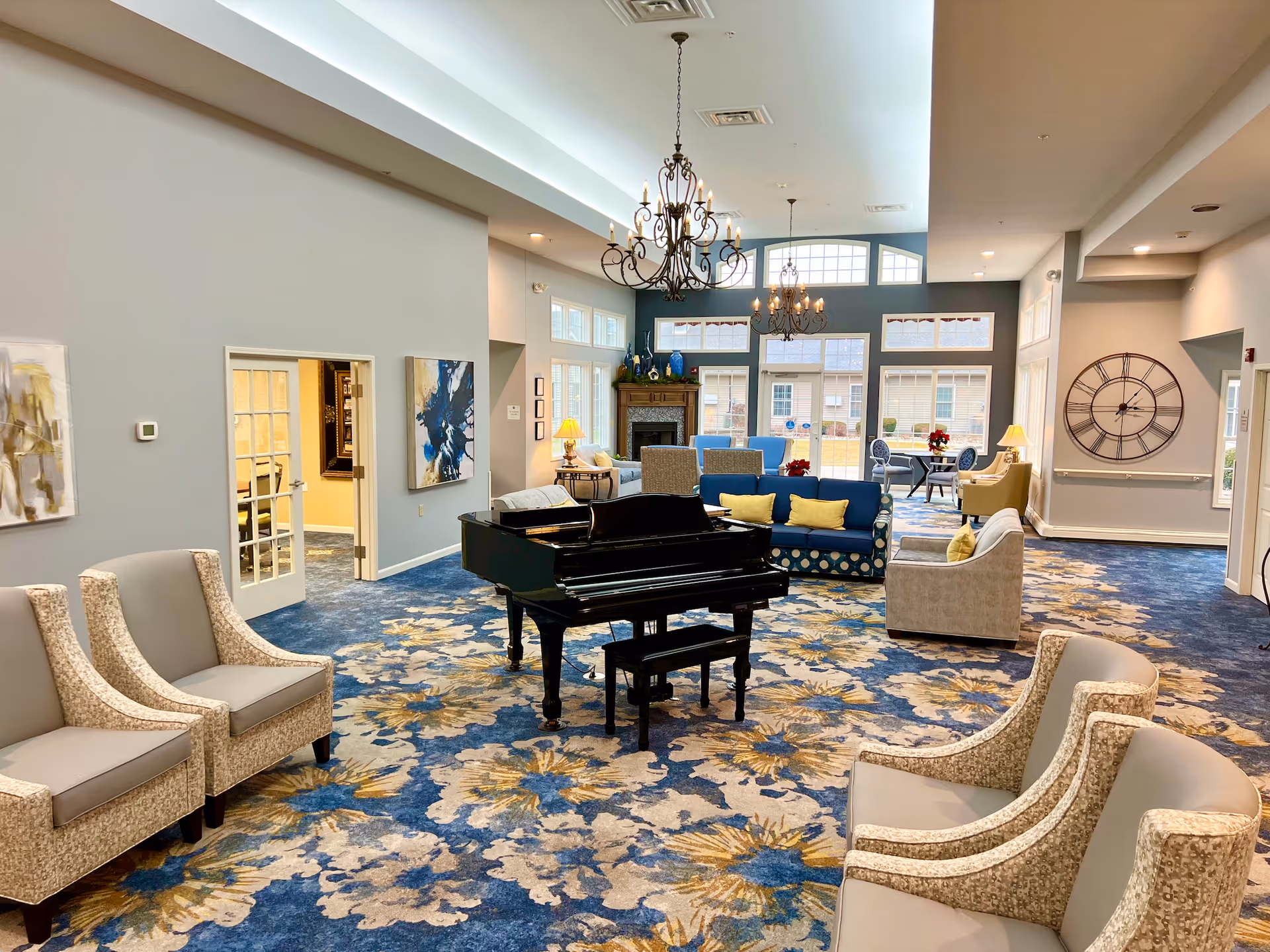 A spacious senior living common area with a floral patterned carpet in blue, beige, and yellow tones. The room features a black grand piano in the center, surrounded by various seating options including beige armchairs, blue sofas with yellow cushions, and small tables with lamps. Large windows and glass doors allow natural light to fill the room, and two ornate chandeliers hang from the ceiling. A large wall clock and decorative artwork are visible on the walls.