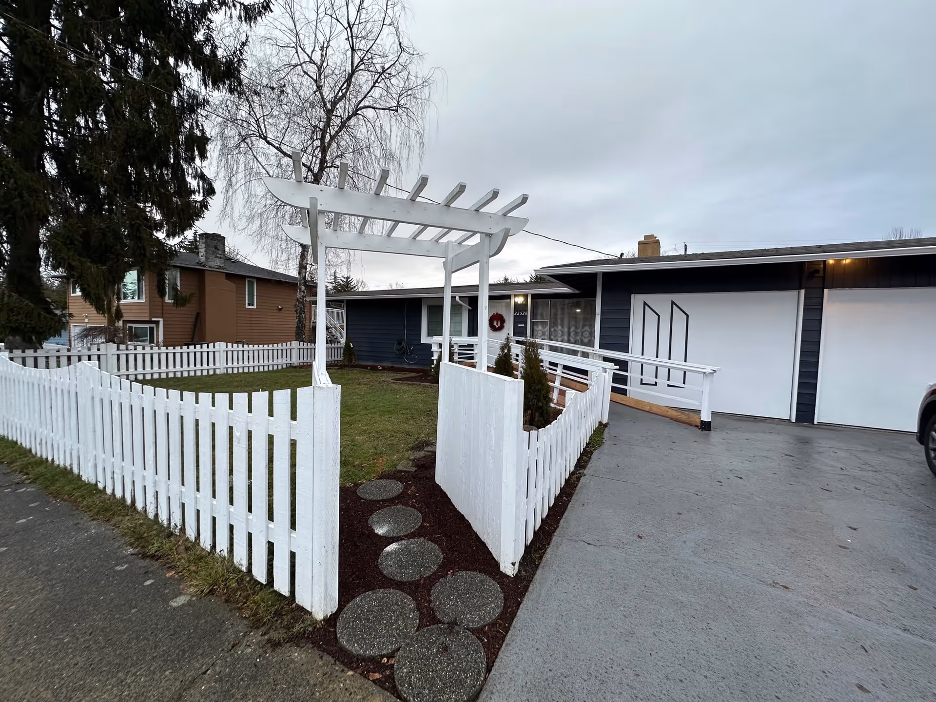 Front exterior of a single-story home with a white picket fence, pergola entrance, small lawn, and attached garage.