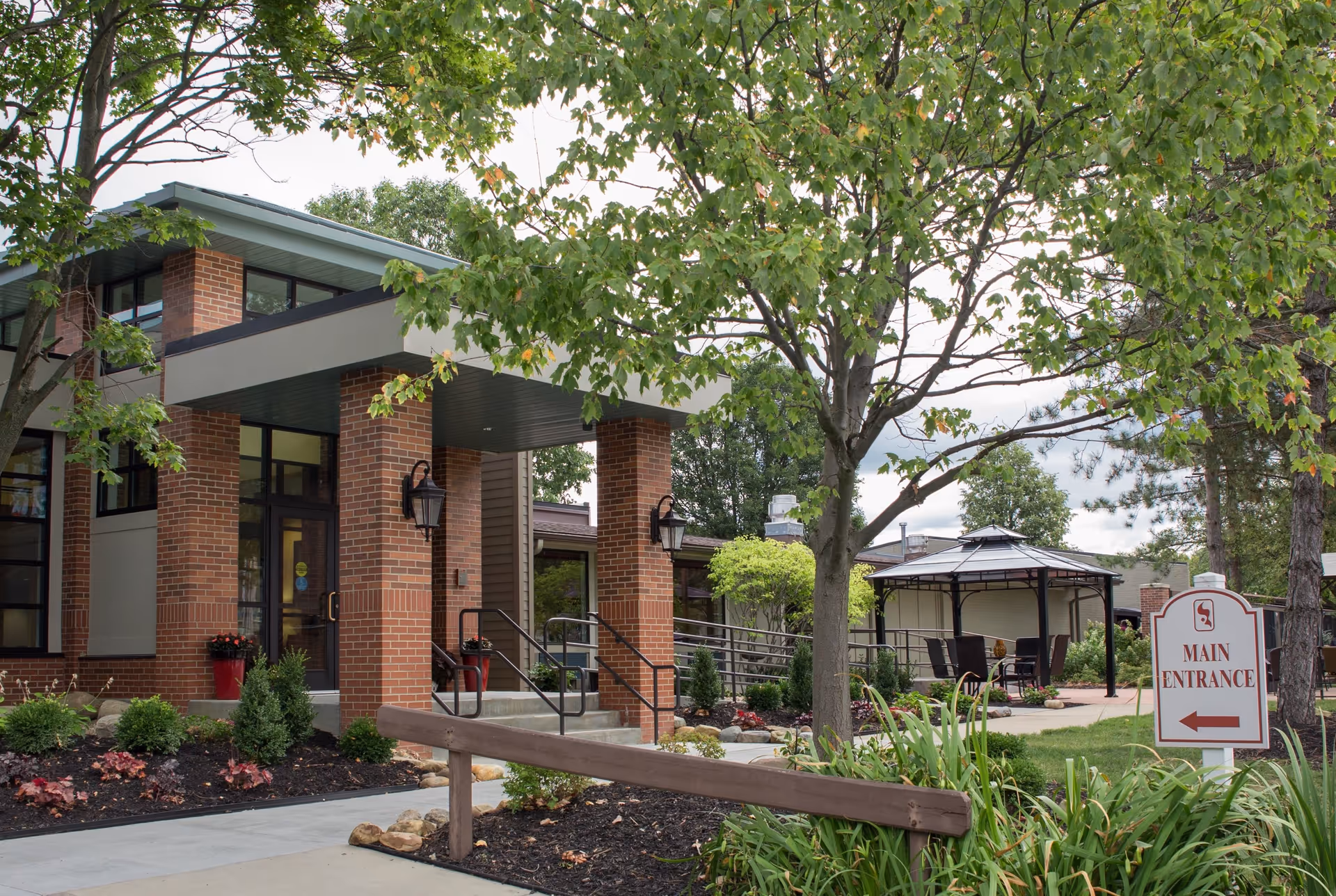 Exterior view of Cardinal Retirement Village showing a brick building entrance with a covered porch, surrounded by landscaped greenery and trees. There is a paved walkway leading to the entrance and a sign indicating the main entrance with an arrow pointing left. A gazebo with outdoor seating is visible in the background.