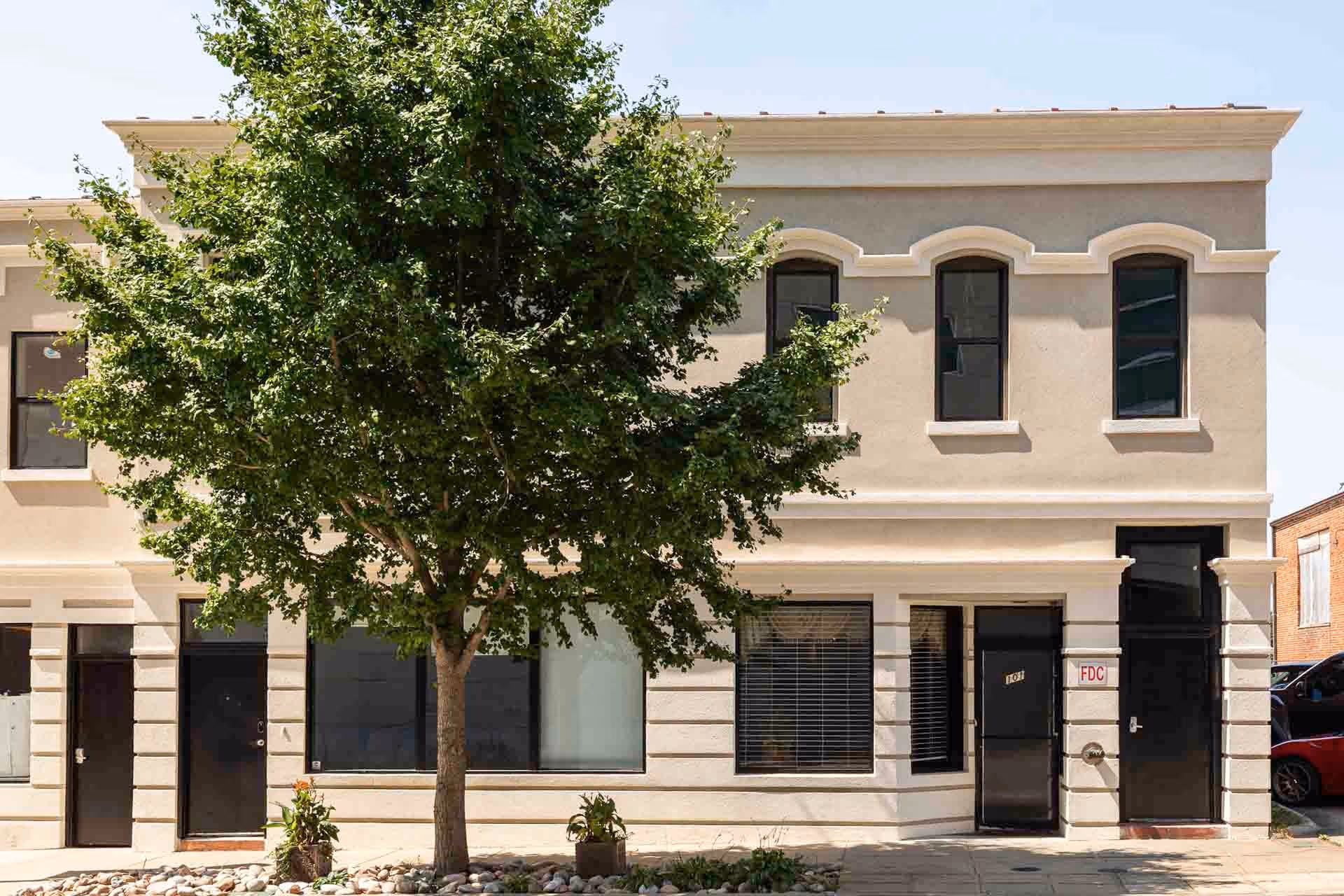 A two-story beige building with arched windows, a tree in front, and a glass door.