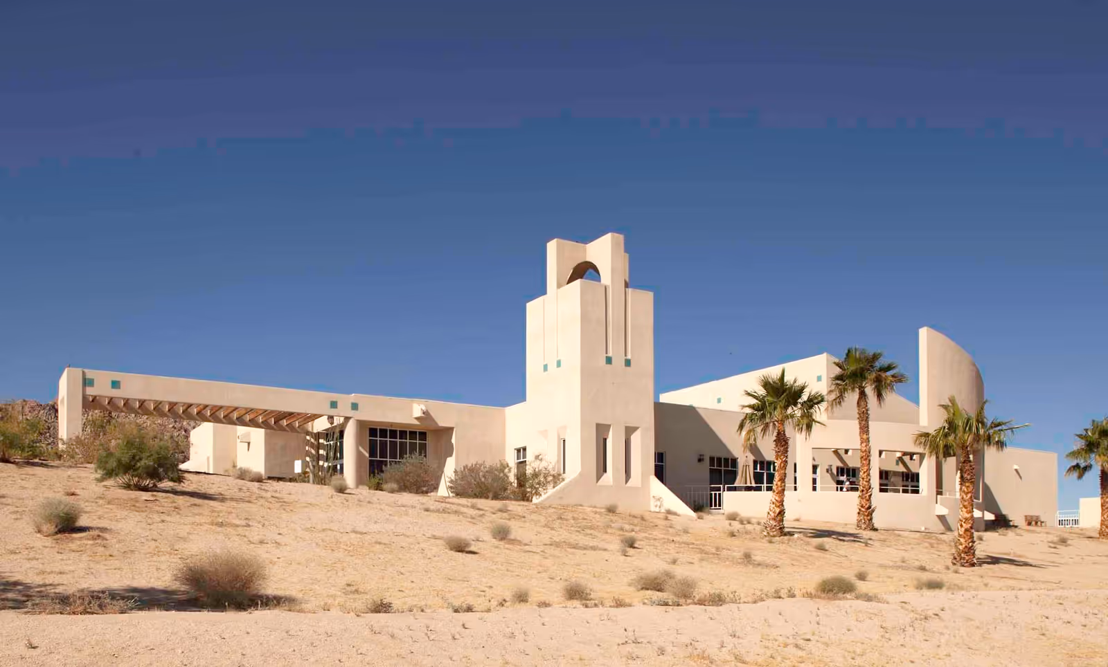 Exterior view of a modern desert-style building with beige stucco walls, a tower-like structure, and several palm trees in front under a clear blue sky.