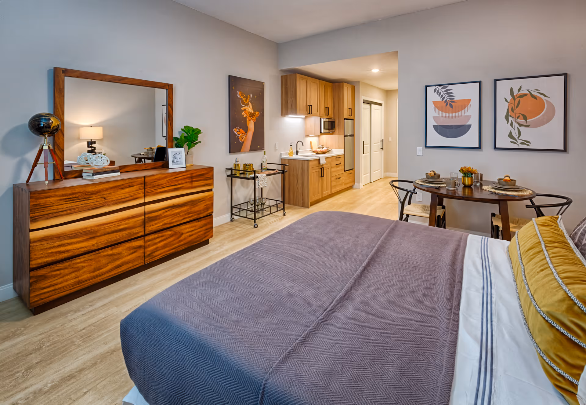 A modern bedroom with a dresser, mirror, table, and kitchenette in the background.