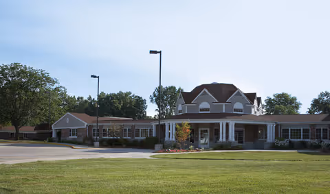 Exterior view of a single-story brick building with a central entrance featuring white columns and a peaked roof, surrounded by a well-maintained lawn and trees under a clear sky.