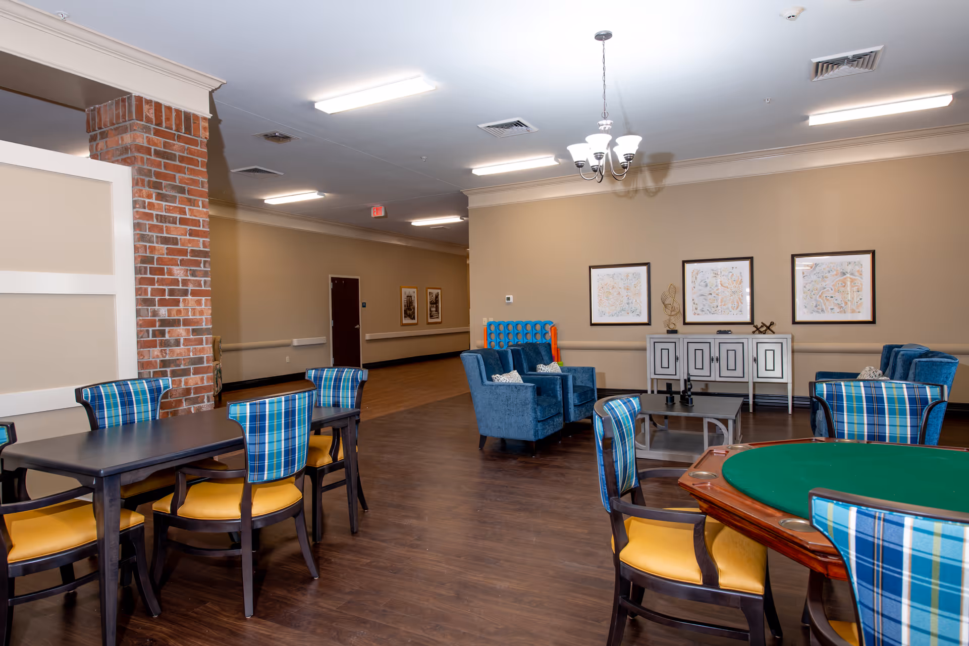 A spacious common area in an assisted living facility featuring multiple seating arrangements including a table with plaid-backed chairs, a green felt card table, and a lounge area with blue armchairs. The room has wood flooring, beige walls, framed artwork, and a brick column.