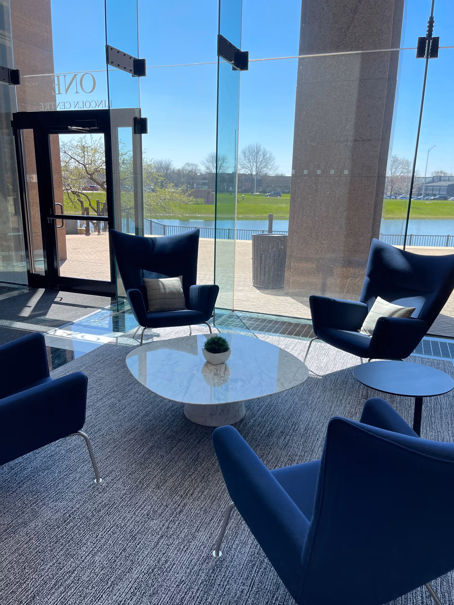 Modern indoor lounge with blue chairs around a round marble coffee table and glass walls.