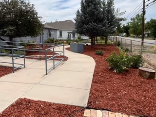 Ramped concrete walkway with metal handrails leads to a building entrance amid red mulch and shrubs.