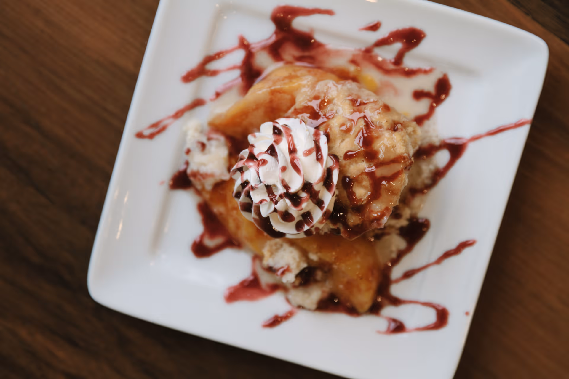 A dessert on a white square plate featuring a pastry topped with whipped cream and drizzled with a red sauce, placed on a wooden table.