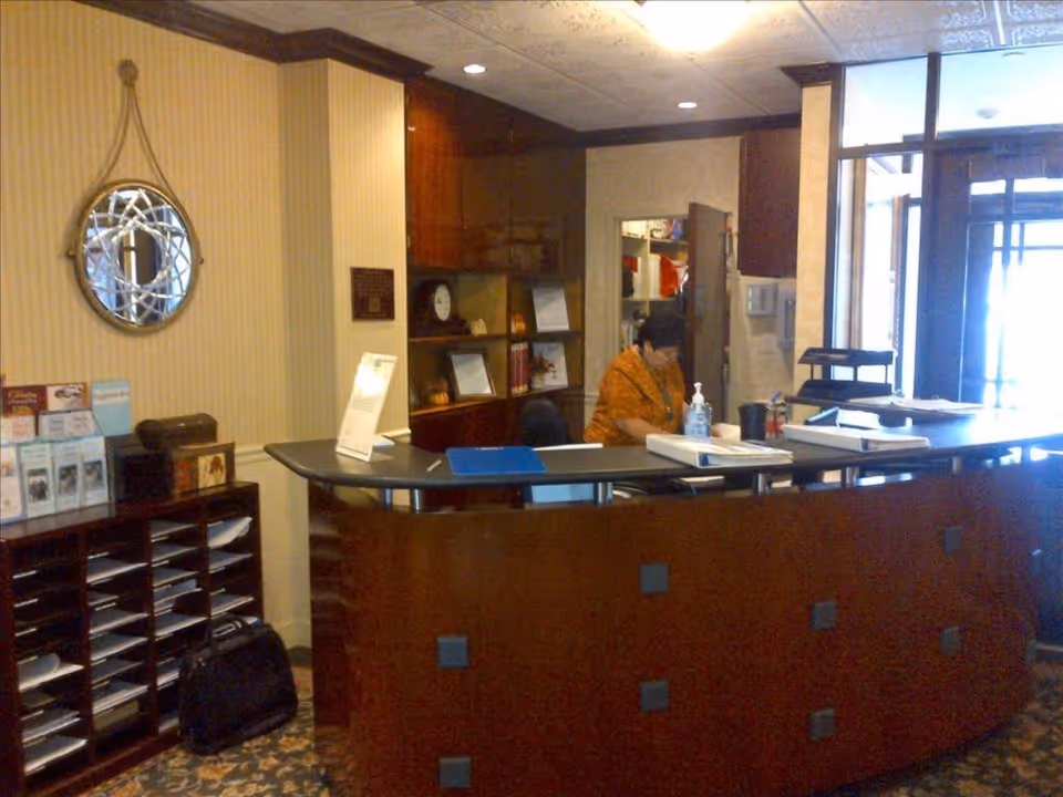 Reception area of a senior living facility with a curved wooden front desk. A person is seated behind the desk working. There are brochures and papers organized on shelves to the left, a decorative round mirror on the wall, and a door leading to another room behind the desk. The area is warmly lit with a patterned carpet and wood trim.