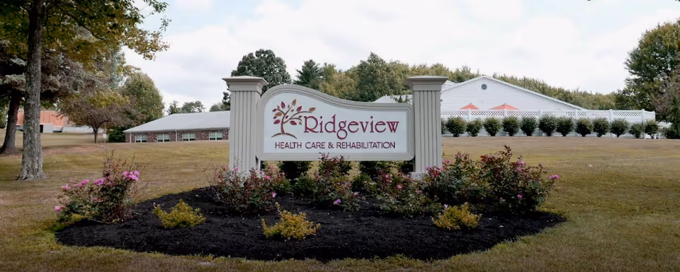 A landscaped outdoor area with a sign that reads 'Ridgeview Health Care & Rehabilitation' surrounded by bushes and flowers, with a building and trees in the background under a partly cloudy sky.