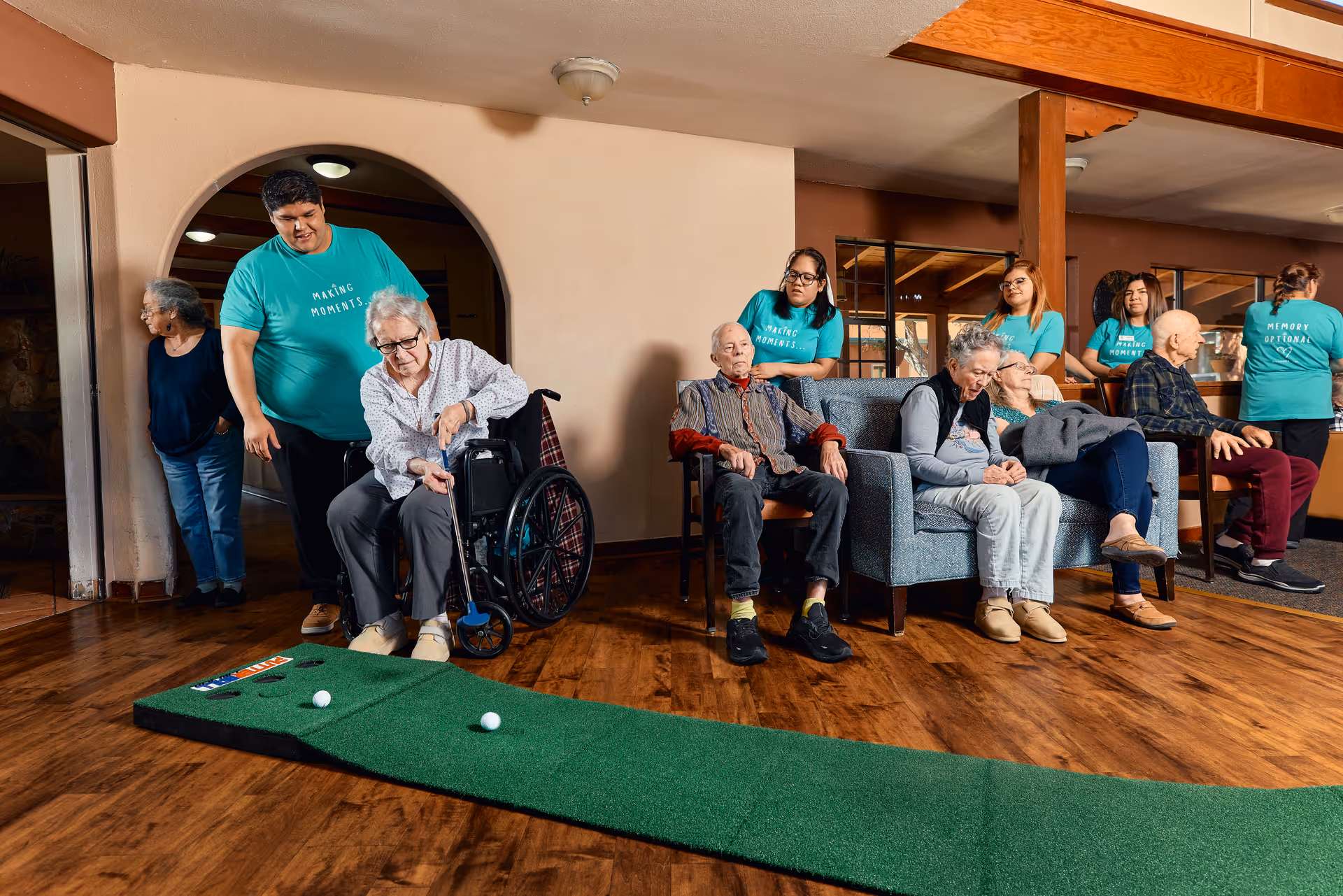 A group of elderly people and caregivers in a senior living facility common area. One elderly woman in a wheelchair is playing indoor mini-golf with assistance from a caregiver wearing a teal shirt that says 'Making Moments.' Other elderly residents are seated on chairs and couches, some interacting with caregivers also wearing teal shirts. The room has wooden flooring and warm lighting.