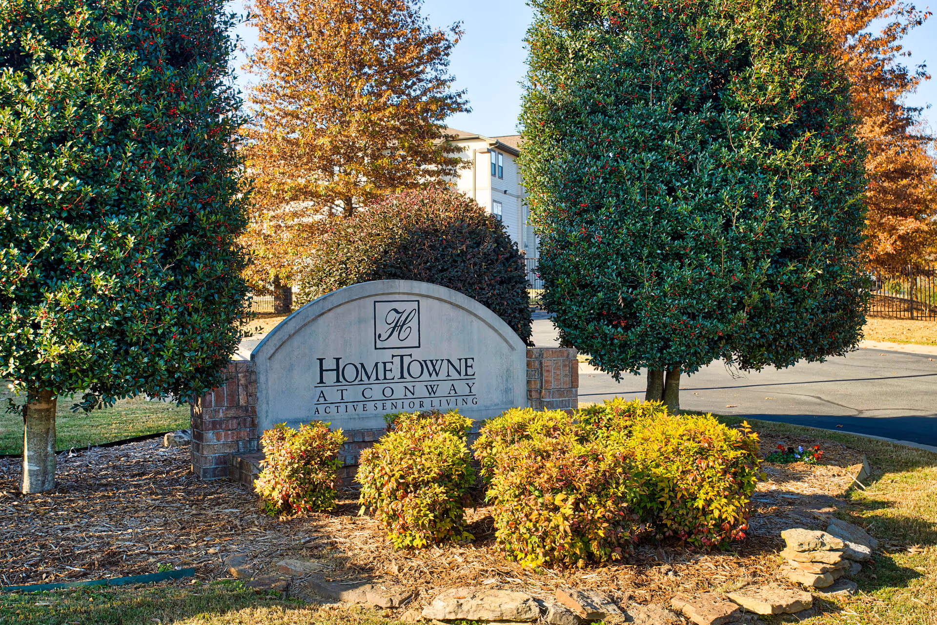 Stone entrance sign reading 'HomeTowne at Conway Active Senior Living' surrounded by landscaped bushes and trees with part of the building visible behind.