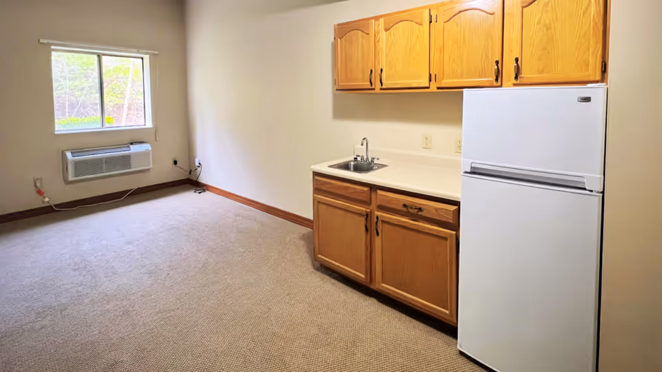 A small kitchenette with wooden cabinets, sink, countertop, and a white refrigerator.