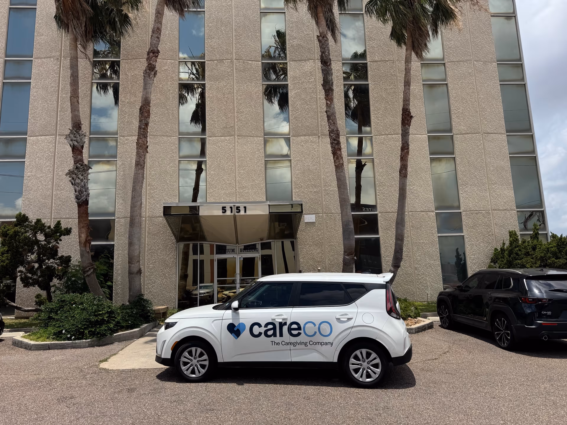 A CareCo-branded car parked in front of a beige multi-story building entrance with palm trees.