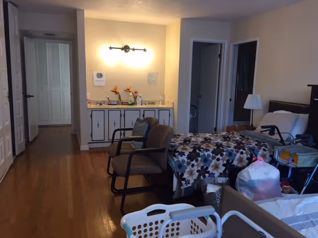 Interior view of a senior living facility room with a bed covered in a floral blanket, two chairs, a small table with a lamp, a walker, and a laundry basket. There is a cabinet with a countertop and flowers against the far wall, and two open doorways leading to other rooms.