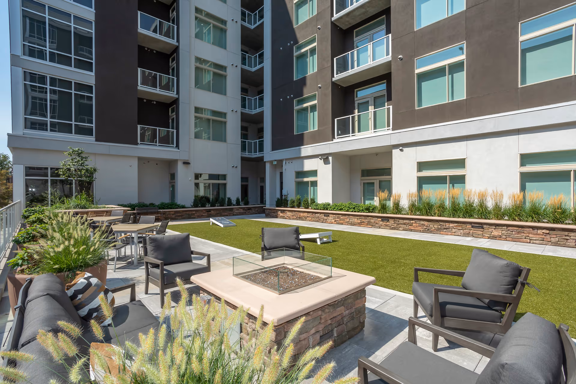 Outdoor patio area at Overture 9th + Co 55+ Apartment Homes featuring cushioned seating around a central fire pit, tables and chairs, decorative plants, and a green lawn with a cornhole game set, surrounded by a multi-story building with balconies and large windows.