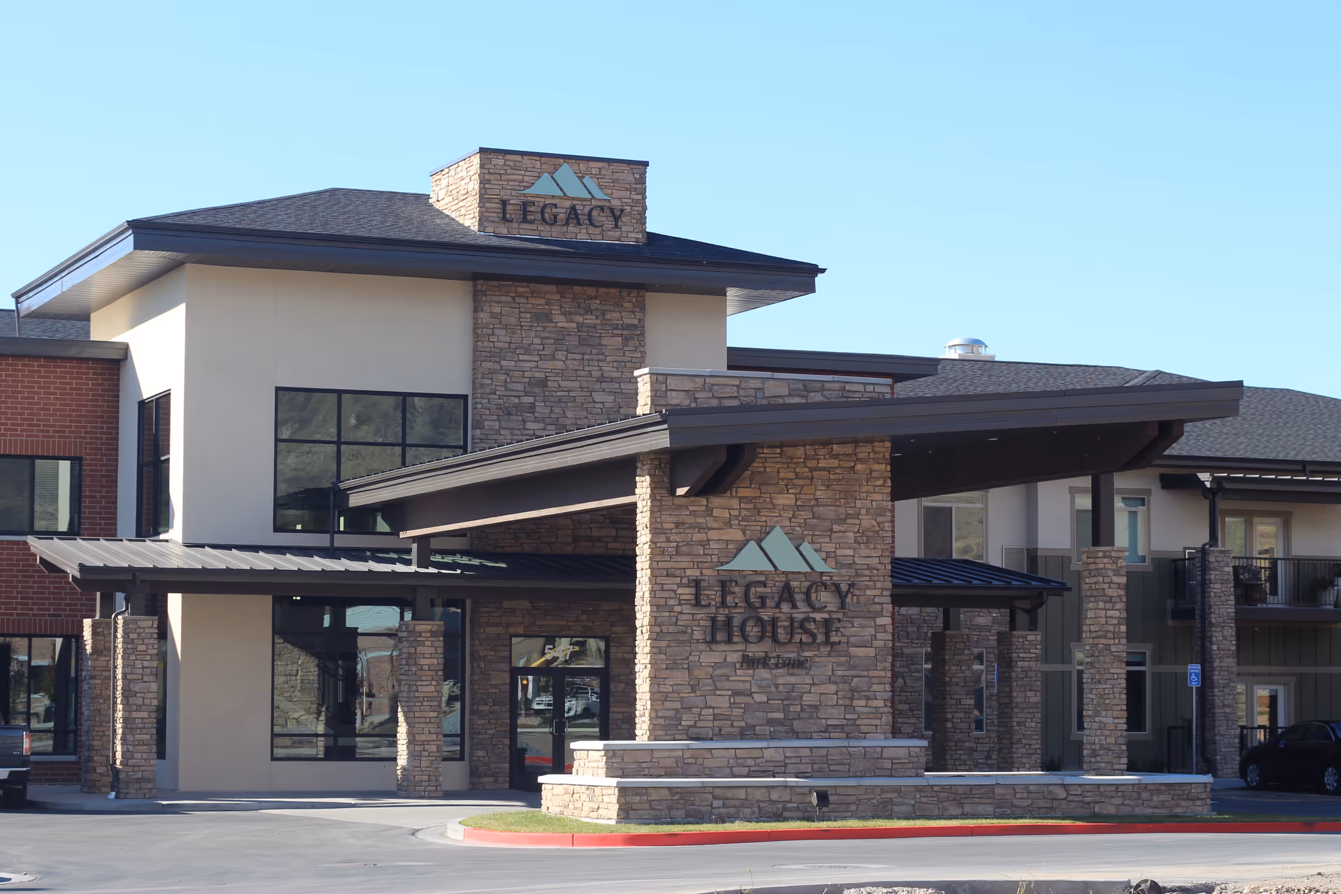 Front exterior view of Legacy House of Park Lane, a senior living facility with stone and beige walls, large windows, and a covered entrance.