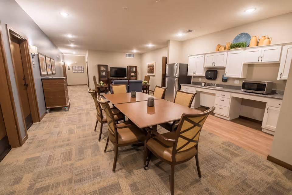 A spacious common area in a senior living facility featuring a dining table with six chairs in the foreground, a kitchenette with white cabinets, a microwave, and a refrigerator on the right, and a seating area with a TV and chairs in the background. The room has beige walls, carpeted flooring, and soft overhead lighting.