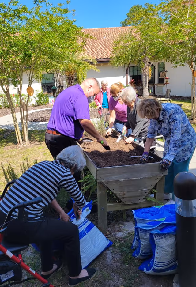 A group of elderly people and a caregiver are gardening outdoors around a raised garden bed filled with soil. They are using small gardening tools and working together under sunny weather with trees and a building with a tiled roof in the background.