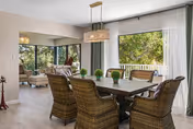 Sunlit dining area with a rectangular table and wicker chairs beneath a pendant light, adjacent to a seating nook and large windows overlooking greenery.