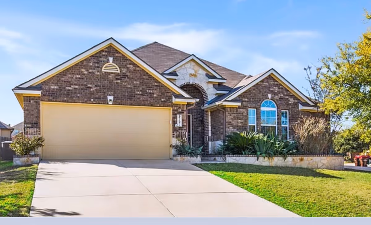Front exterior view of a single-story brick house with a large beige garage door, a concrete driveway, and a well-maintained lawn with shrubs and trees under a clear blue sky.