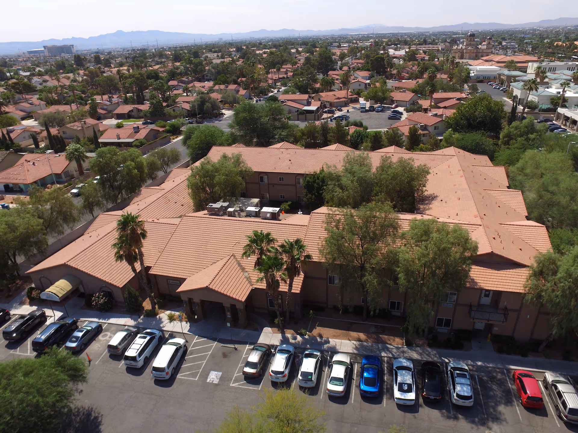 Aerial view of Sana Assisted Living facility showing a large building with a red-tiled roof surrounded by trees and a parking lot with several cars parked. The surrounding neighborhood with many houses and distant mountains is visible in the background.