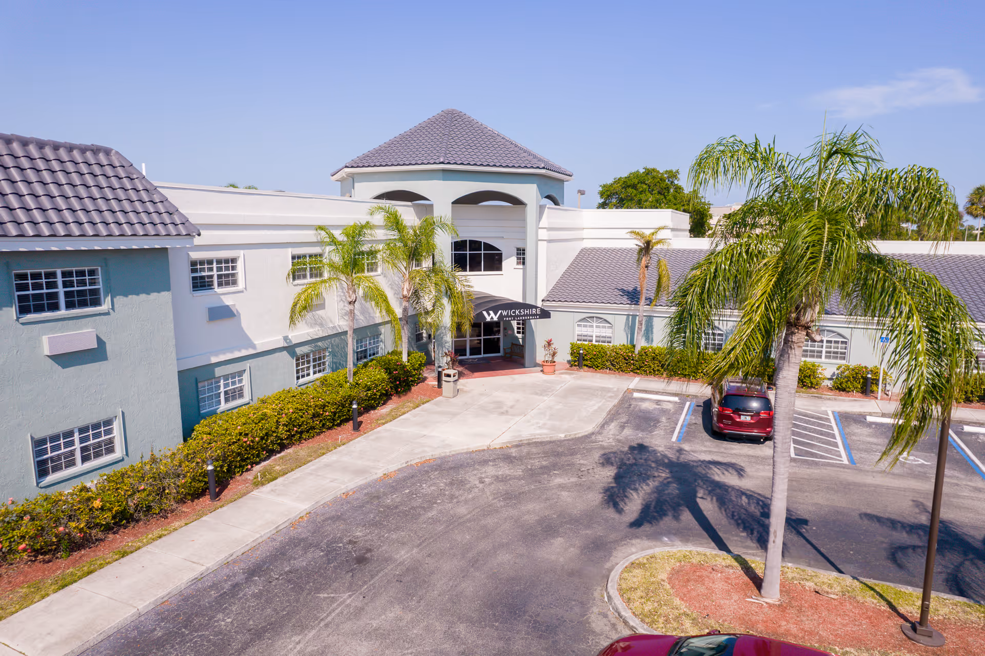 Exterior view of a senior living facility named Wickshire with a light blue and white building, palm trees, and a parking lot with a few cars under a clear blue sky.