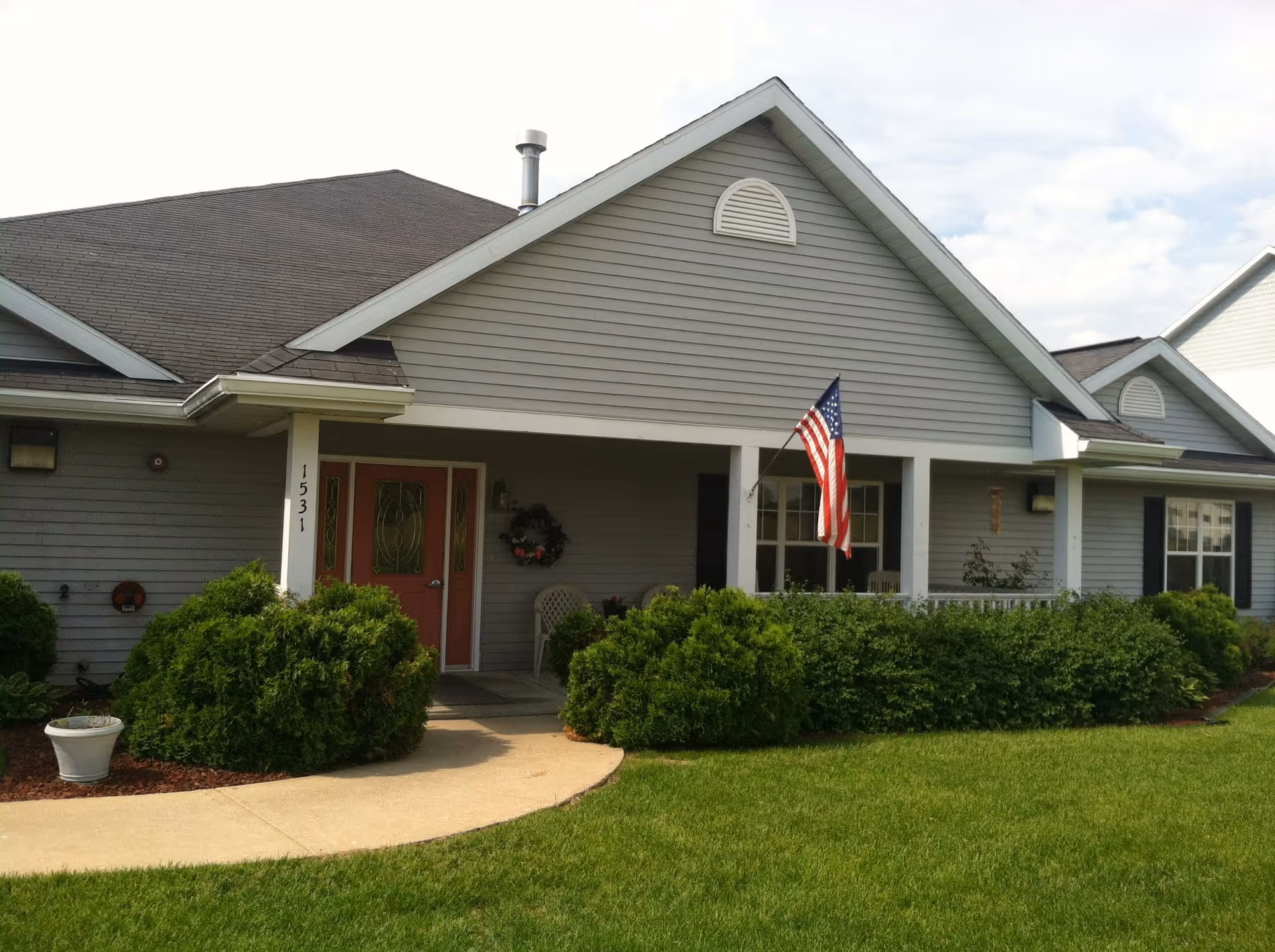 Exterior view of a single-story building with gray siding and a dark roof. The entrance features a covered porch with white columns, a red door with decorative glass panels, an American flag mounted on a pole, and green bushes lining the walkway and front of the building. The building number 1531 is visible on a column near the door.