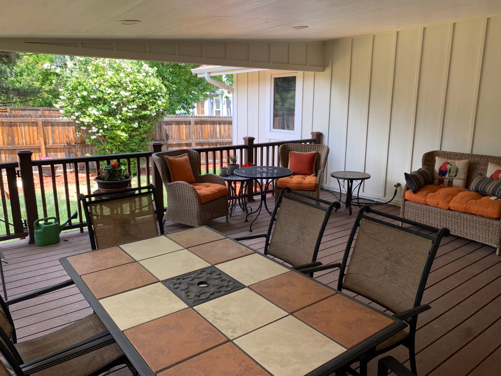 Covered outdoor patio area with a tiled table and four chairs in the foreground. In the background, there are two cushioned wicker armchairs with orange cushions and a small round table between them, as well as a cushioned wicker loveseat with decorative pillows. The patio overlooks a fenced backyard with green grass and a flowering bush.