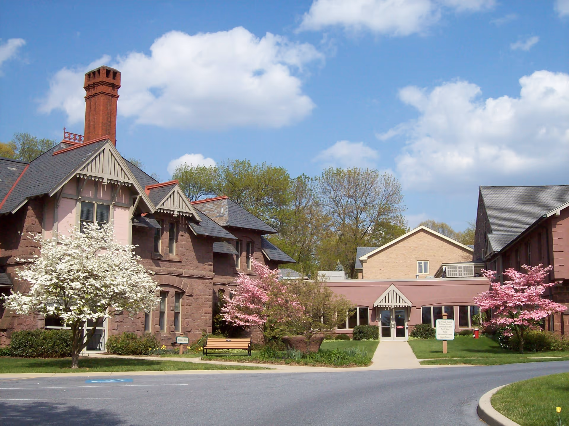 Front view of a brick senior living building with flowering trees, a bench, and a central entrance under a blue sky.