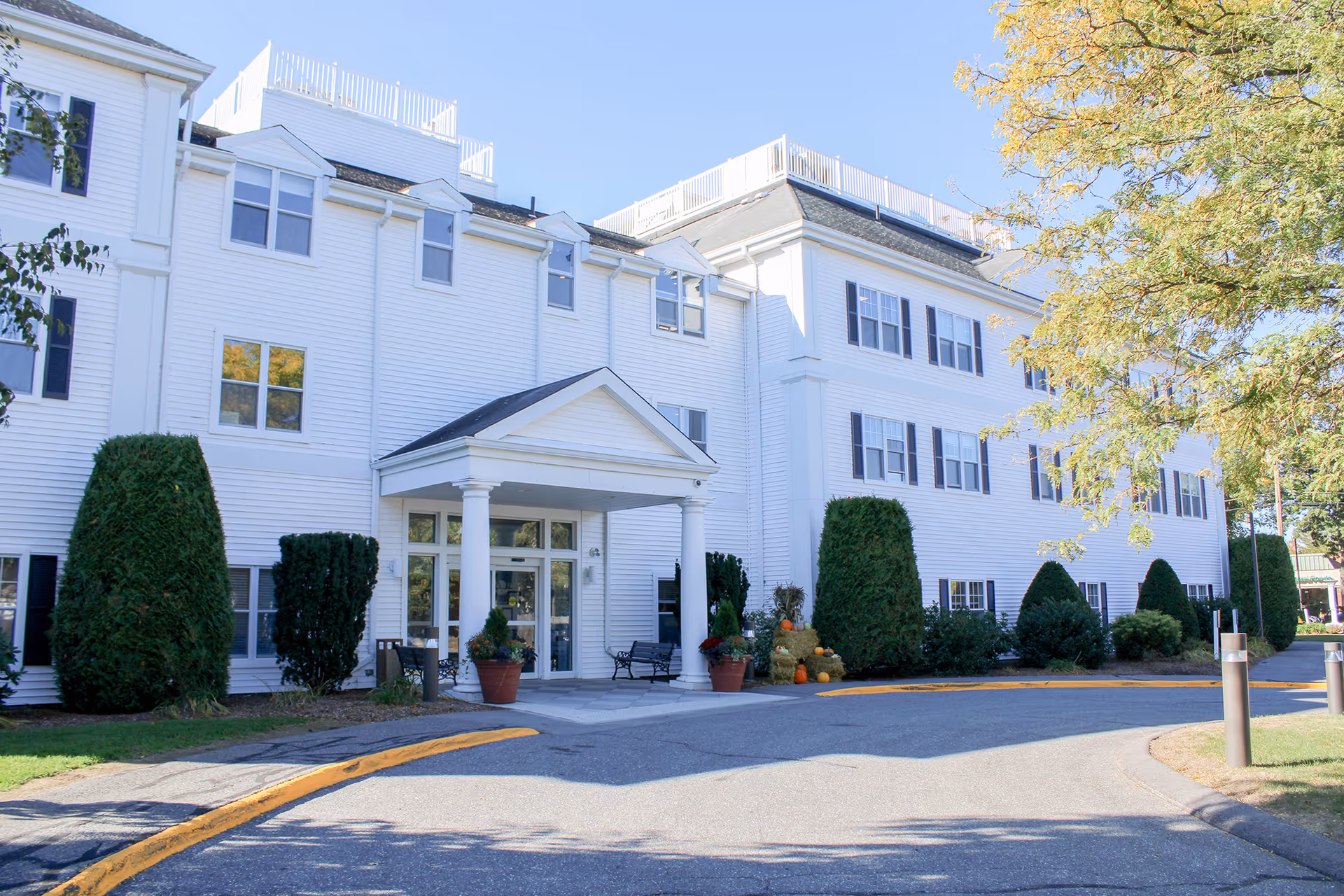 White multi-story senior living building with a front entry, manicured shrubs, and pumpkins.