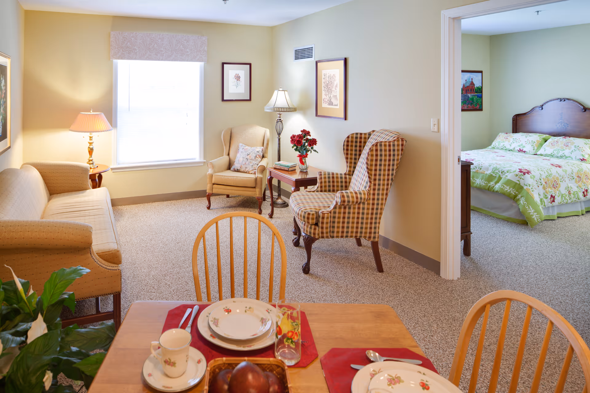 Well-lit sitting area with upholstered chairs, a sofa and a dining table, opening into a bedroom with a floral bedspread.