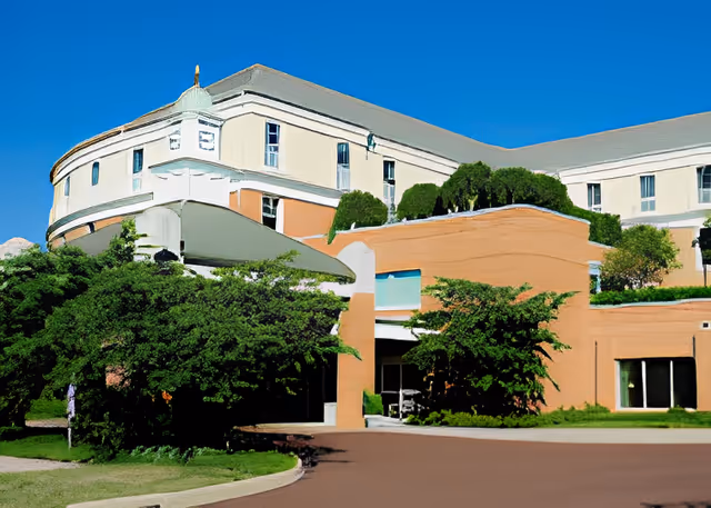 Front exterior of a multi-story senior living facility with curved architecture, landscaping, and a driveway.