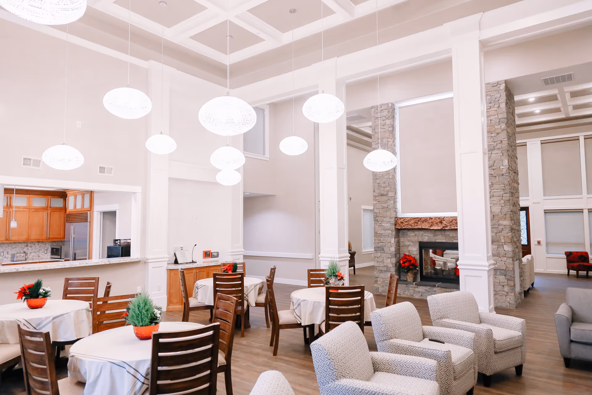 Bright senior living dining area with round tables, chairs, white walls, and a stone fireplace.